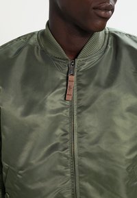 Olive green bomber jacket, made from smooth, shiny fabric. Features a zip front with a leather pull tag and ribbed collar and cuffs.