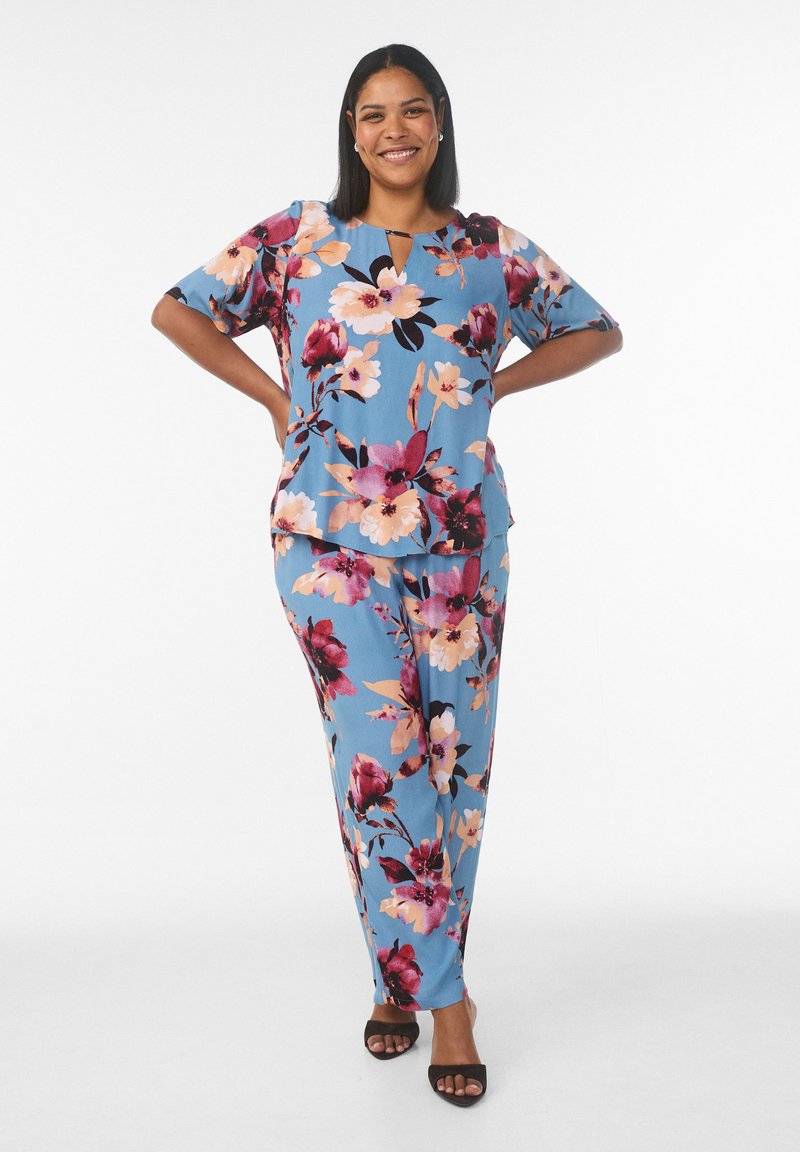 Blaugrünes Outfit mit Blumenmuster aus leichtem Stoff; kurzärmeliges Oberteil mit Schlitz am Ausschnitt, passende weit geschnittene Hose. Schwarze Sandalen.