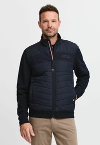 Marineblaue Reißverschlussjacke mit gepolstertem Vorderteil und einfarbigen Ärmel, ausgestattet mit einem Stehkragen und Logodetail. Glatte Textur und elastische Bündchen.