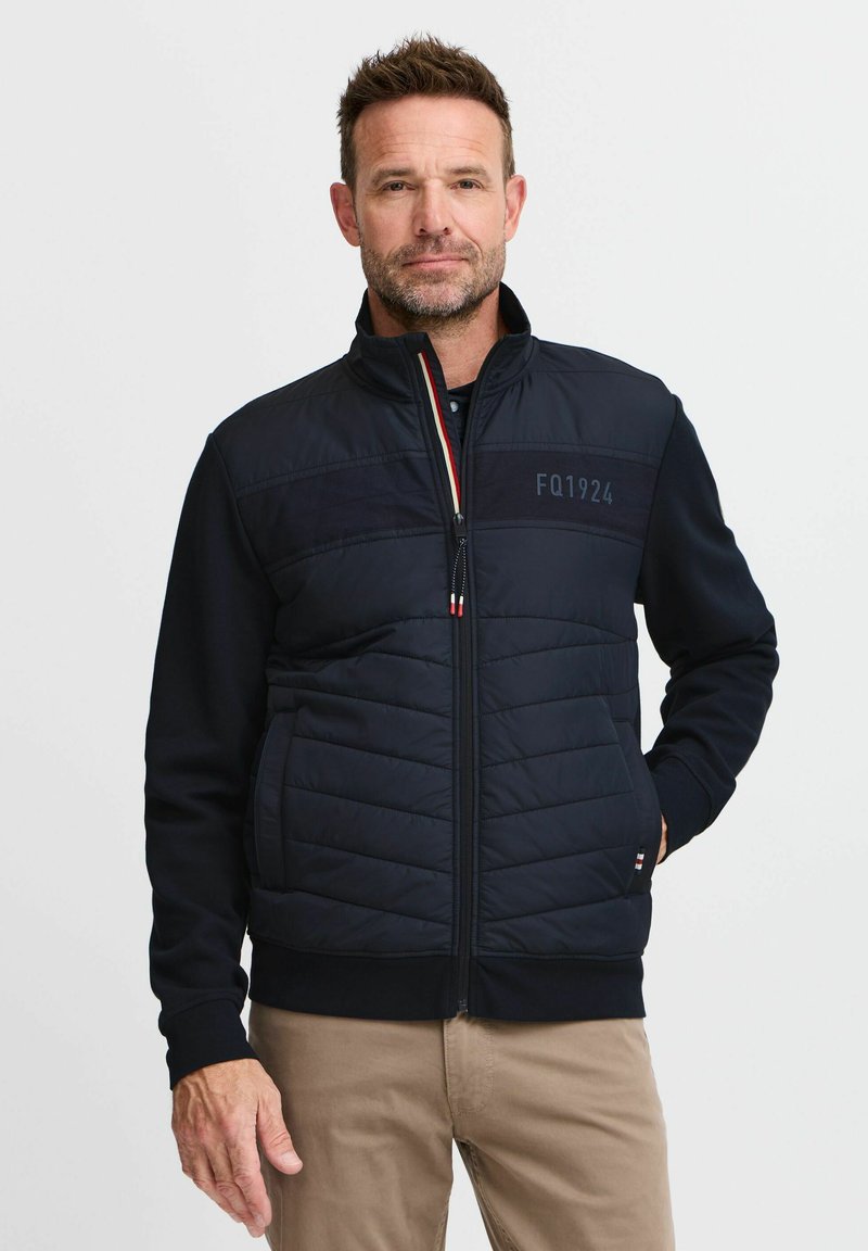Marineblaue Reißverschlussjacke mit gepolstertem Vorderteil und einfarbigen Ärmel, ausgestattet mit einem Stehkragen und Logodetail. Glatte Textur und elastische Bündchen.