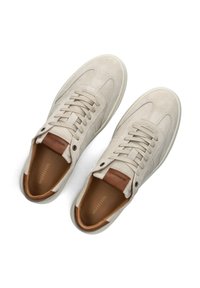 Beige suède sneakers met een witte rubberen zool, met een bruine leren accent op de hak en platte veters door de voorste lus.