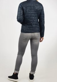 Granatowa kurtka puchowa o pikowanym wzorze, szare dopasowane dżinsy oraz czarne botki za kostkę z białymi podeszwami, widoczne z tyłu na białym tle.