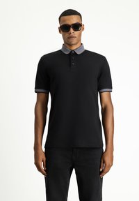 Schwarzes Polohemd mit grauem Kragen und Akzenten an den Ärmeln, mit einer Drei-Knopf-Leiste und einem klassischen Schnitt. Kombiniert mit schwarzen Jeans.