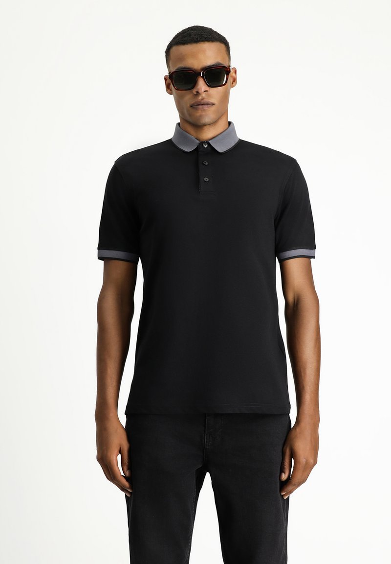 Schwarzes Polohemd mit grauem Kragen und Akzenten an den Ärmeln, mit einer Drei-Knopf-Leiste und einem klassischen Schnitt. Kombiniert mit schwarzen Jeans.