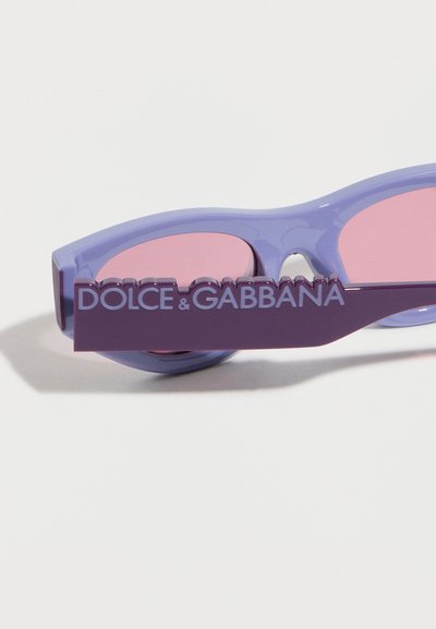 Μωβ γυαλιά ηλίου με ροζ φακούς και ανάγλυφο λογότυπο "Dolce & Gabbana" στον βραχίονα, σε λευκό φόντο.