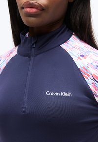 Κοντινό πλάνο γυναίκας που φοράει μπλε ναυτικό τοπ Calvin Klein με φερμουάρ στον γιακά και πολύχρωμες, σχεδιασμένες πάνελ στους ώμους.