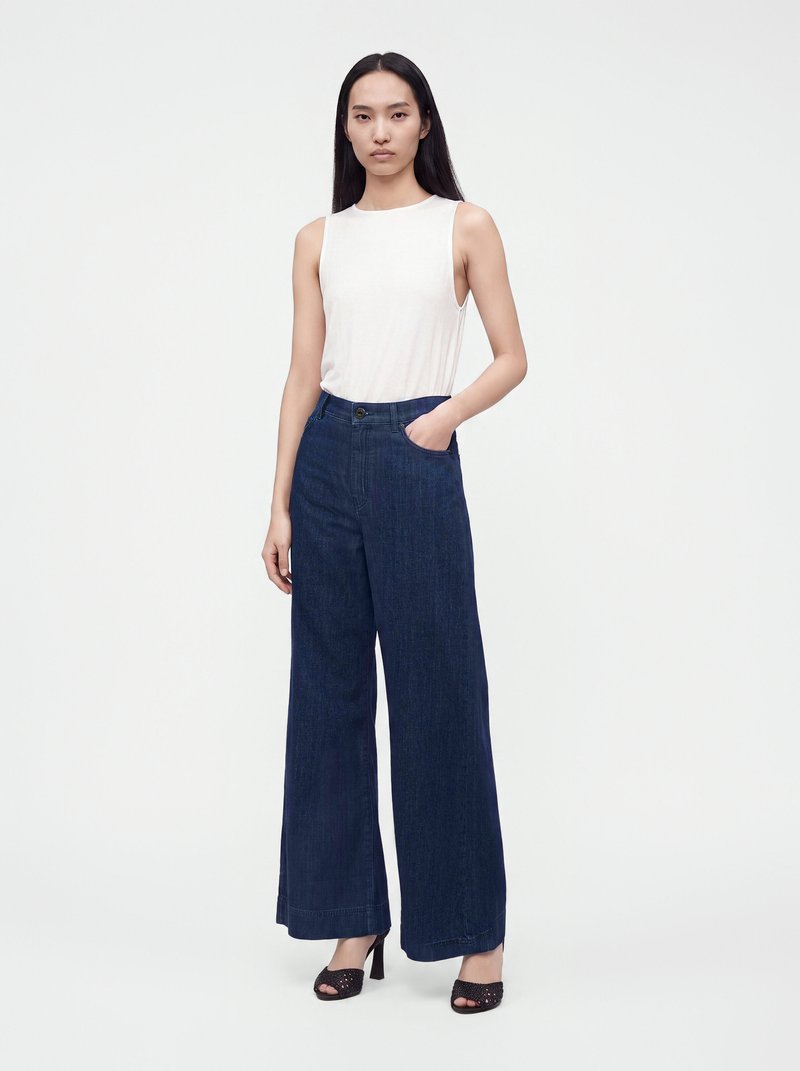 Woman with long black hair wearing a sleeveless white top, dark wide-leg jeans, and black open-toe heels, standing with one hand in pocket.