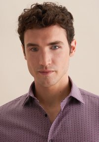 Koszulka z krótkim rękawem w kolorze bordowym z geometrycznym wzorem, z kołnierzykiem zapinanym na guziki i gładką teksturą materiału. Zbliżenie na twarz modela.