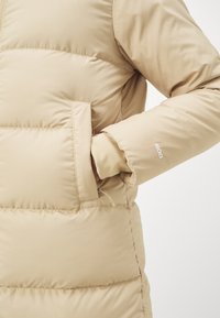 Giubbotto imbottito beige con zip frontale, tasca laterale e design a trama imbottita. Presenta polsini elastici e il marchio "600" sulla manica.