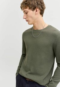 Young man wearing a textured olive green sweater, silver chain necklace, and dark trousers, looking downwards against a plain light background.