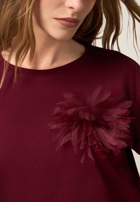 T-shirt bordeaux avec un appliqué floral doux et superposé sur la poitrine, featuring des pétales transparents et une texture de tissu lisse.
