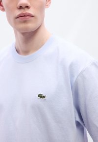 Ανοιχτό μοβ βαμβακερό μπλουζάκι με στρογγυλή λαιμόκοψη και κεντητό πράσινο λογότυπο κροκόδειλου στο αριστερ ό στήθος. Λείο ύφασμα και κλασική γραμμή.