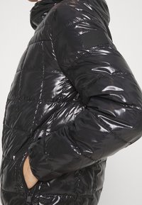 Homme portant une veste matelassée noire brillante avec une poche zippée et un patch logo Calvin Klein sur la manche supérieure, les mains dans les poches.