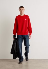 Rød crewneck sweatshirt i et glat stof, parret med mørkeblå jeans. Modellen holder en sort læderjakke i den ene hånd.