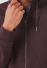 Haut de sweat à capuche zippé de couleur bordeaux foncé, avec une texture lisse, des poignets côtelés et un logo brodé discret sur la poitrine gauche.