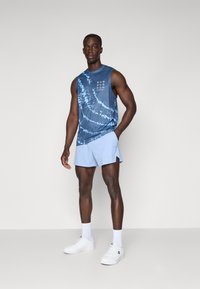 Débardeur de sport en motif bleu tie-dye, associé à un short bleu clair. Le modèle porte des baskets blanches et des chaussettes, mettant en valeur le design de vêtements de sport.