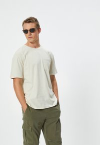 Beige bomulls-t-shirt med rund hals och korta ärmar; har en bröstficka. Bärs med olivgröna cargobyxor, händerna i fickorna.