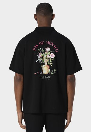 Mann trägt schwarzes Kurzarmshirt mit pinkem Blumenstrauß und Text "PAS DE MONACO" und "FLORALE À FLEUR DE PEAU" auf der Rückseite.