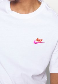 Bílá bavlněná tričko s kulatým výstřihem a krátkými rukávy. Na levé straně hrudi se nachází malá vyšívaná značka Nike v růžové a oranžové barvě.