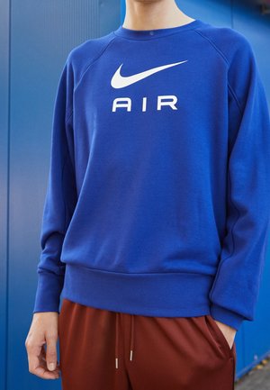 Osoba ubrana w niebieską bluzę Nike Air i brązowe spodnie, stojąca na tle niebieskiej ściany, ręka w kieszeni.