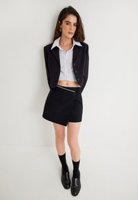 Zwarte cropped blazer over een wit gestreept shirt, gecombineerd met zwarte shorts met witte biezen, zwarte knie hoge sokken en zwarte loafers.