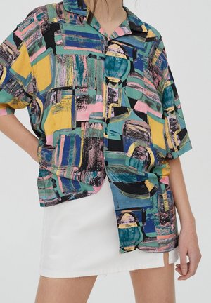 Femme portant une chemise boutonnée à motifs abstraits multicolores à manches courtes et une mini-jupe blanche.