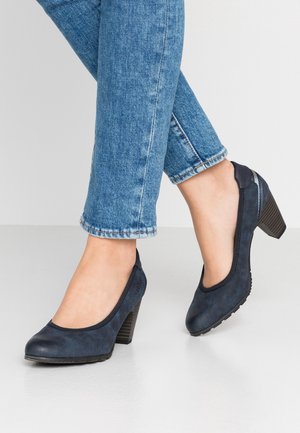 Ben iført blå jeans og marineblå lukkede sko med lav hæl, der står på en hvid gulv mod en ensfarvet lys baggrund.