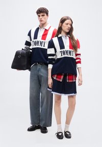 Νέος άνδρας και γυναίκα που φορούν ταιριαστά πουλόβερ Tommy με ρίγες σε κόκκινο, λευκό και μπλε, στέκονται μπροστά σε ένα απλό λευκό φόντο.