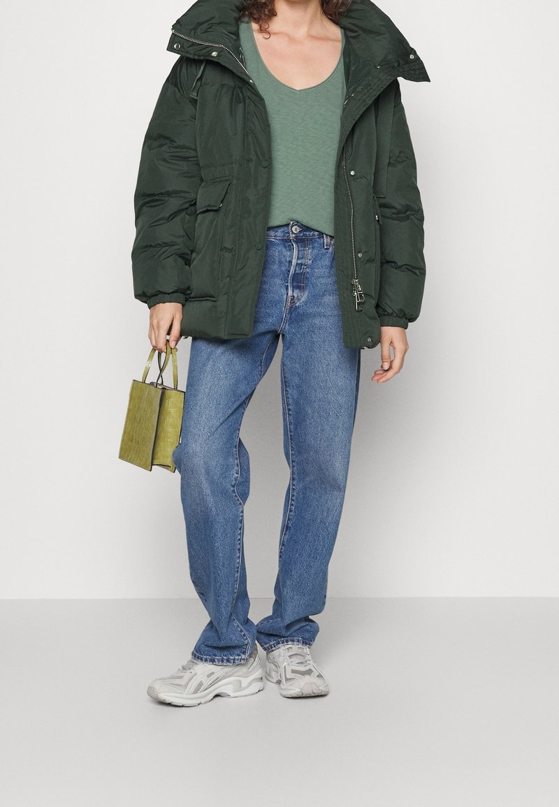 Personne portant une doudoune vert foncé, un haut vert, un jean bleu, des baskets blanches et tenant un petit sac à main vert.