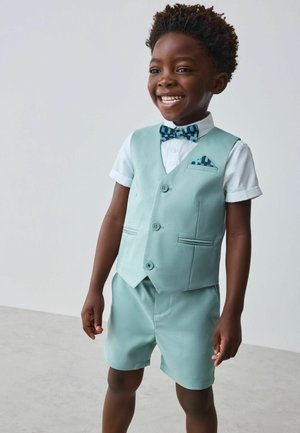 Usmievavý chlapec oblečený v mintovo zelenom veste a kraťasoch s bielou košeľou, modrým kockovaným motýlikom a zodpovedajúcou vreckovkou v saku stojaci v interiéri.