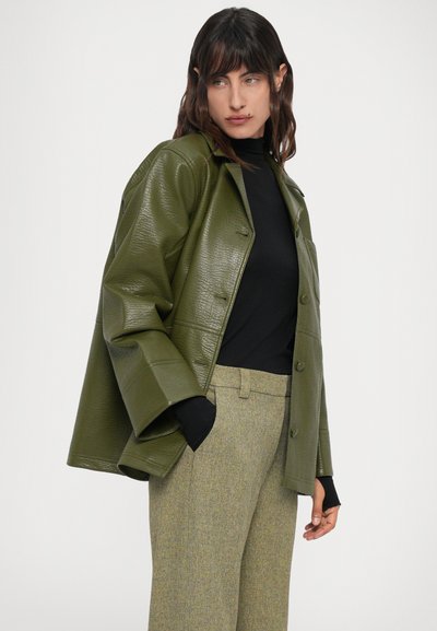 Donna che indossa un giubbotto di pelle oversize verde oliva sopra una maglietta nera e pantaloni grigi chiari, in piedi con le mani in tasca.