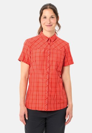 Lächelnde Frau mit braunen, zurückgebundenen Haaren, die ein rotes karierte Kurzarm-Hemd mit Knöpfen und schwarze Hosen vor weißem Hintergrund trägt.