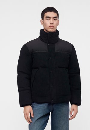 Homme portant une doudoune noire avec un col montant et un jean bleu, debout devant un fond clair uni.