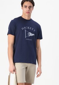 Camiseta de algodón azul marino con un logo gráfico plateado que presenta un diseño de bandera. Se lleva con pantalones cortos beige, sostenida con un bolso de color claro.