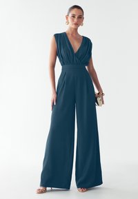 Teal värvi kombinesoon sügava V-kaelusega, laiad jalad ja kogutud vöökoht. Sile kude ja ilma varrukateta disain. Mudel hoiab käes kuldset kotikest.