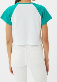 Bílý zkrácený tričko s térovými raglánovými rukávy. Měkká látka, krátké rukávy a lem, který končí nad pasem. V kombinaci s bílými kalhotami.