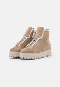 Bottines en daim beige avec une doublure en peluche, présentant une semelle épaisse blanche et un bout arrondi. Lacets et accents de couture visibles.