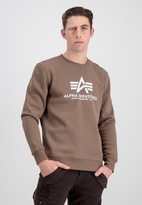 Sudadera marrón con cuello redondo, que presenta un estampado de logo blanco y puños acanalados sutiles. Tejido texturizado con un bolsillo lateral visible.