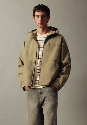 Jeune homme portant une veste à capuche beige par-dessus une chemise blanche et noire rayée avec un jean gris délavé, debout les mains dans les poches.