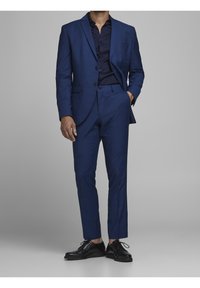 Tmavě modrý oblek na míru s odpovídajícími slim-fit kalhotami a černými společenskými botami. Košile pod ním je tmavě modrá a má hladkou strukturu.