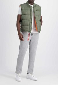 Grüne Steppweste mit orangefarbenem Innenfutter, ausgestattet mit einer Fronttasche, kombiniert mit grauen Jogginghosen und weißen Sneakers. Mit einem roten Etikett-Detail.