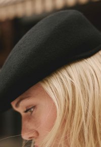 Schwarzer Filzhut mit glatter, abgerundeter Form, der teilweise das hellblonde Haar bedeckt. Nahaufnahme betont Textur und Kanten.