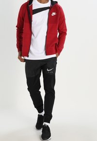 Červená mikina na zip s černými akcenty, bílá tričko s horizontálním černým pruhem, černé tepláky s šedým logem Nike a černé tenisky.