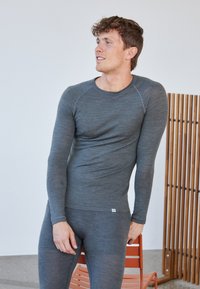 Mand i tætsiddende grå langærmet trøje og leggings, læner sig op ad en orange stol og kigger sidelæns mod en ensfarvet hvid væg i baggrunden.