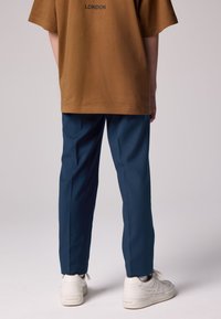 Ruskea lyhythihainen paita, jossa "LONDON"-teksti; tummansiniset suoralinjaiset housut; valkoiset tennarit teksturoiduilla yksityiskohdilla.