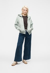 Νεαρή γυναίκα που στέκεται φορώντας μια ανοιχτοπράσινη μπλούζα με κουκούλα GAP, μαύρο t-shirt, φαρδιά μπλε τζιν και μπεζ αθλητικά παπούτσια με σχέδια σε λευκό φόντο.