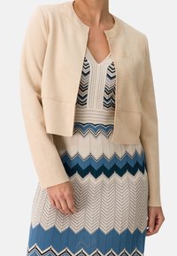 Chaqueta corta de ante sintético beige con cuello redondo y sin cierres, llevada sobre un vestido estampado en tonos azules y crema.