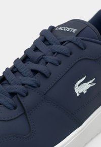 Marineblaue Sneaker aus glattem synthetischen Material, mit Schnürsenkeln und einem weißen Krokodil-Logo an der Seite. Gummisohle sichtbar.