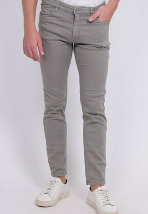 Homme portant un pantalon gris slim et des baskets blanches, debout avec une main dans la poche contre un fond clair uni.