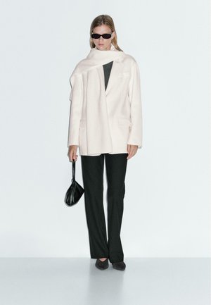 Manteau oversize rose clair avec poches, porté sur un haut sombre, associé à un pantalon noir et un petit sac à main noir. Le mannequin porte également des lunettes de soleil.